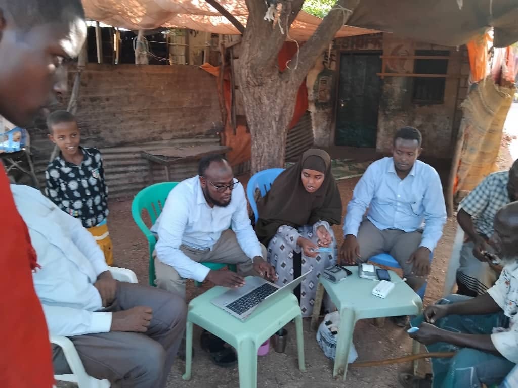 Men and a woman seated on plastic chairs under a tree, gathered around low tables with a laptop and handheld devices, a child watching from the side