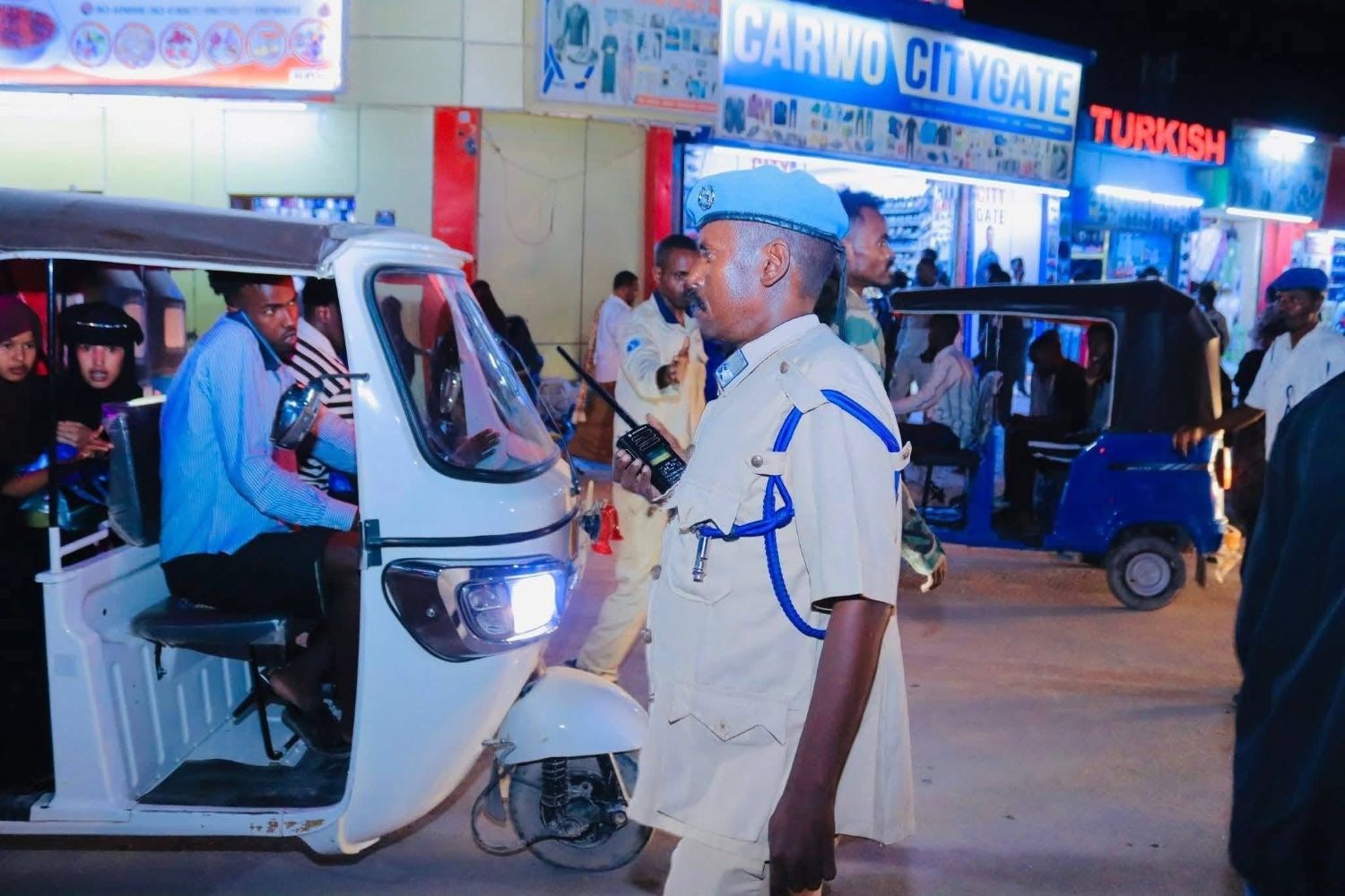 A traffic police officer in a khaki uniform and blue beret, with a whistle on a blue lanyard and a radio in his hand, directs a bajaj full of passengers outside lit shopfronts at night in Las Anod