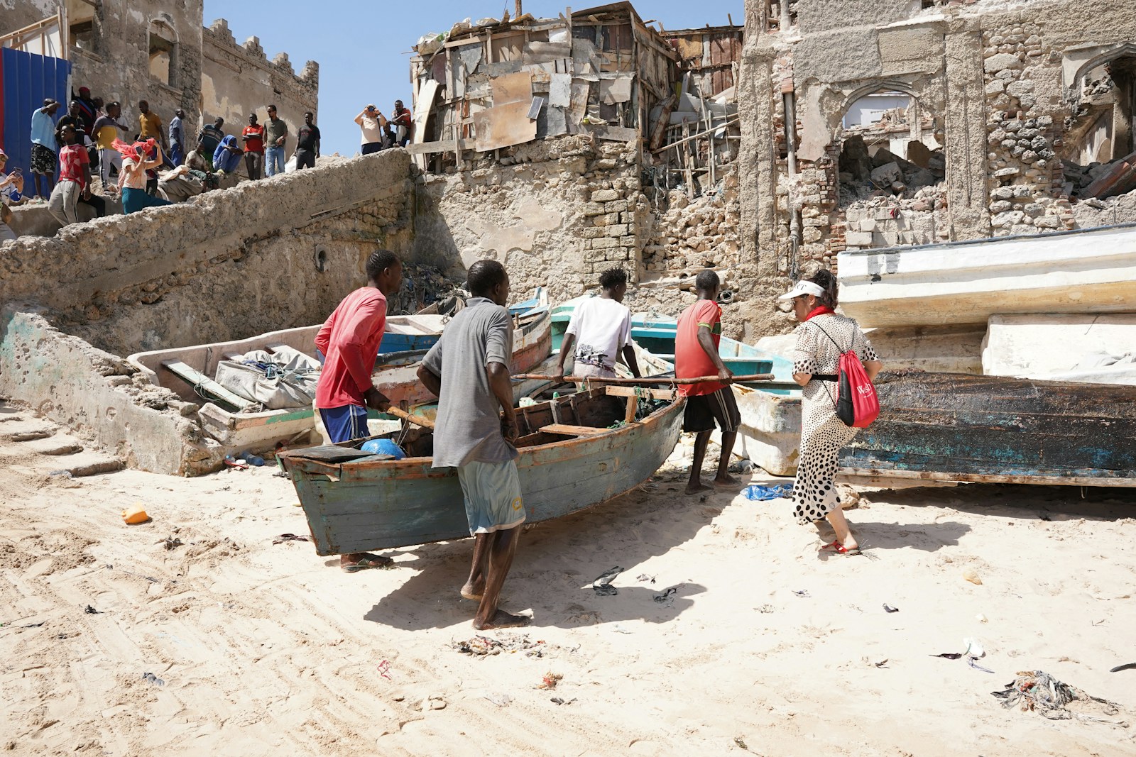 Fishermen haul a weathered wooden fishing boat up a sandy beach by hand, with old stone buildings behind them